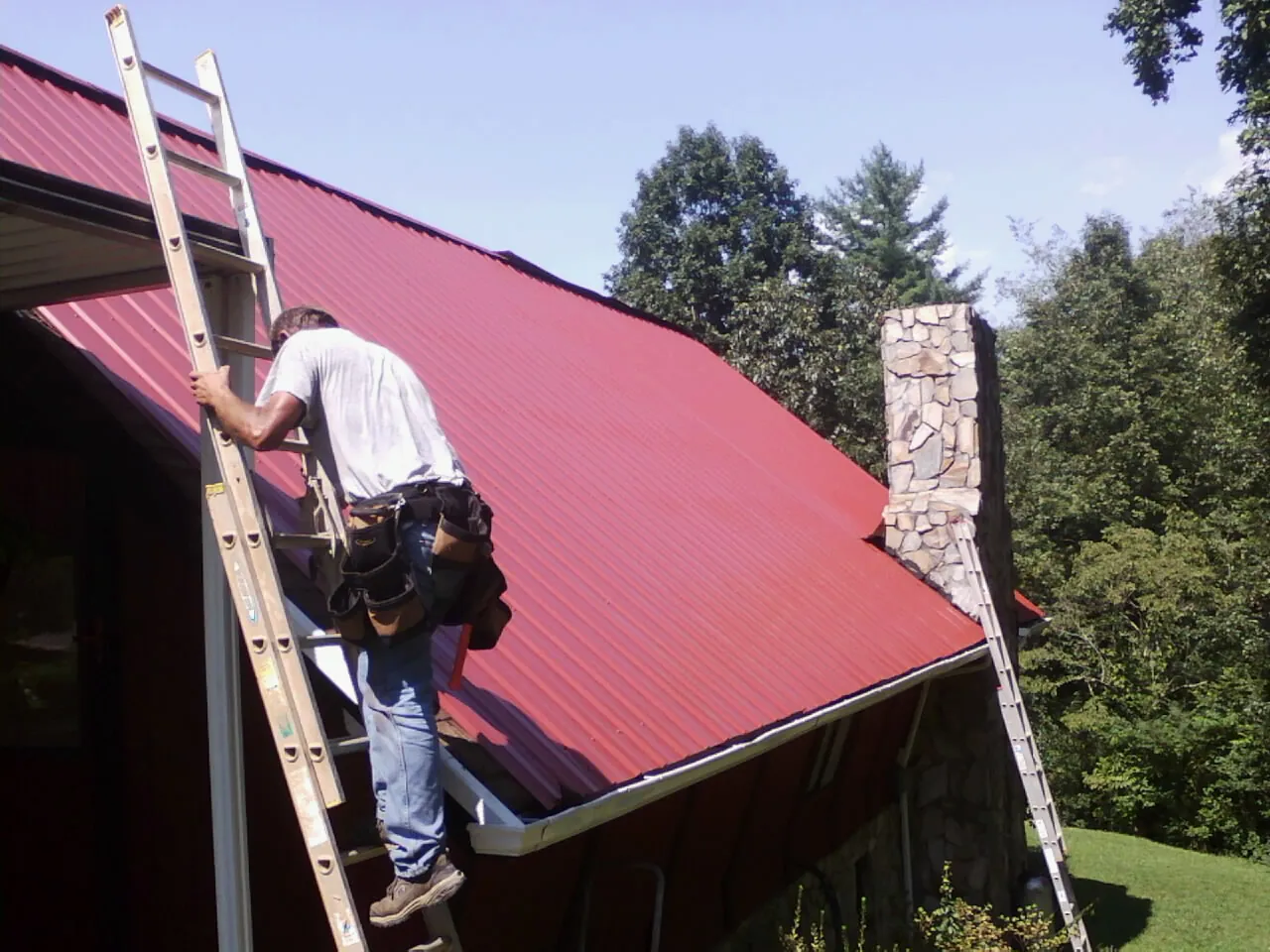 Roofer on a ladder beside a red metal roof and stone chimney — Soffit & Fascia Repair by Roofing Brothers in Aultman